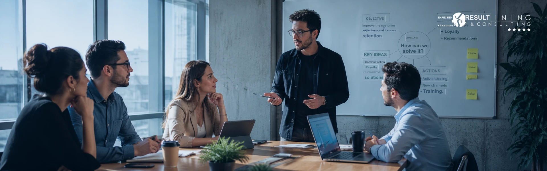 A team in a meeting room listening to a facilitator in front of a board with diagrams and notes.