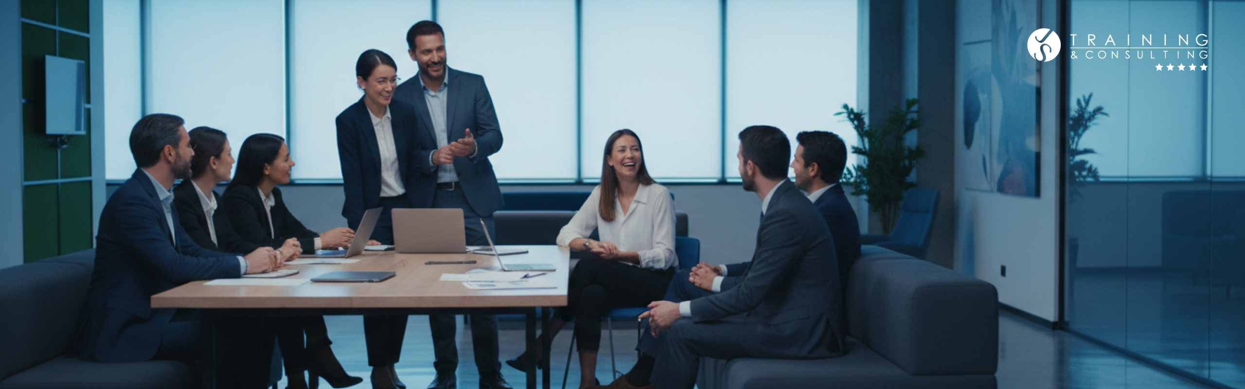 Equipo de profesionales en una reunión corporativa alrededor de una mesa con ordenadores portátiles, sonriendo y colaborando en una oficina moderna.